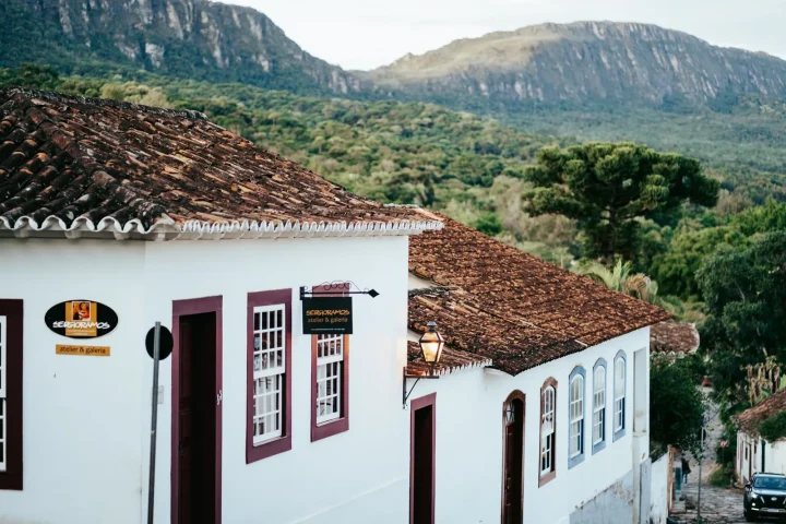 Casarão colonial de paredes brancas e telhado de telha com montanhas ao fundo